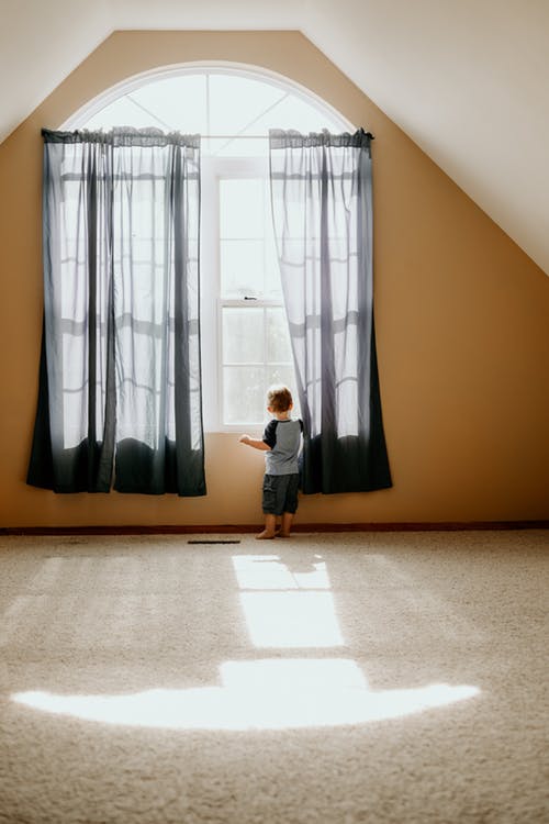 Little boy in a carpeted room looking out the window; there are several FAQ for home sellers that should be addressed before selling.