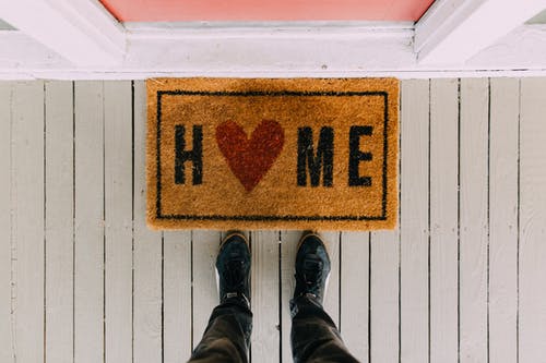 A person's feet standing at the front door of a home with a welcome mat that says home.